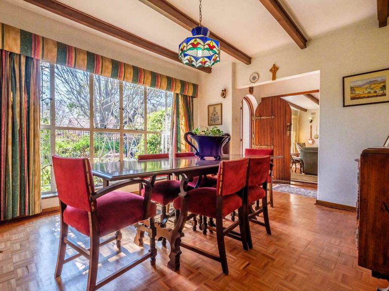 A traditional dining room featuring parquet flooring, exposed timber ceiling beams, and a large window overlooking the garden, with an arched entrance to the living area.