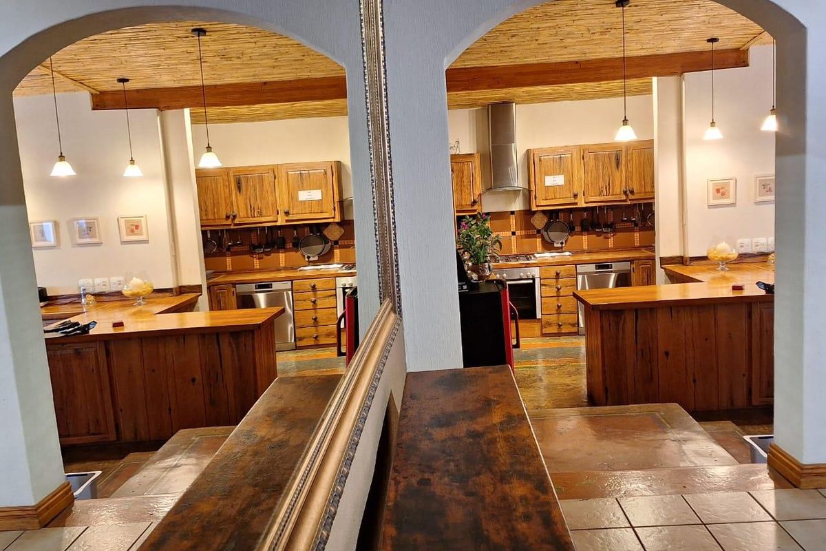 A rustic-style kitchen featuring wooden cabinetry, a tiled backsplash, integrated appliances, and a distinctive timber-slat ceiling, viewed through decorative arched openings.