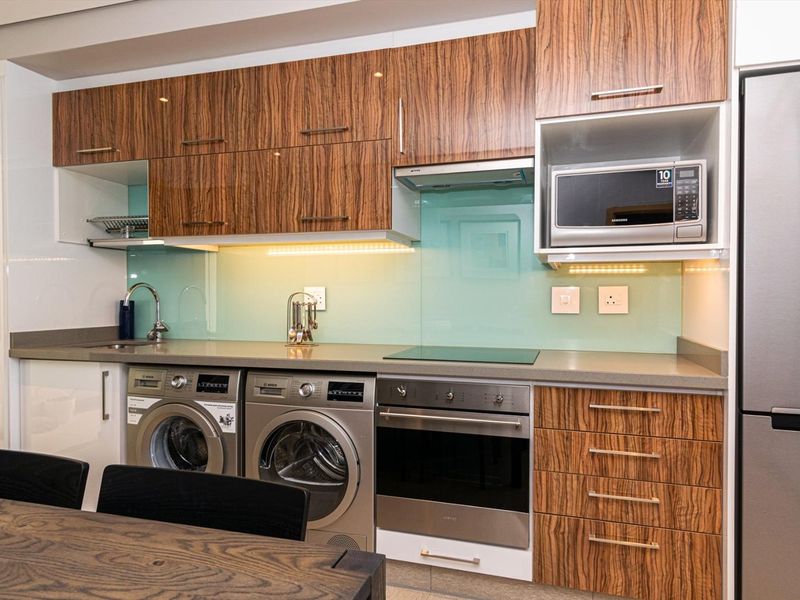 Modern kitchen featuring wood-grain cabinetry, stone countertops, glass splashback, and integrated appliances including an oven, hob, and microwave.