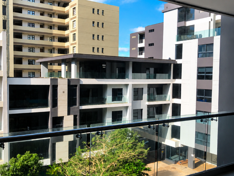 A modern covered balcony featuring a sleek glass balustrade and tiled flooring, offering an elevated view of the surrounding apartment complex.