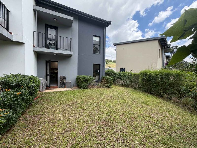 Rear view of a modern townhouse featuring a private garden, paved patio area, and an upper-level balcony with metal railings.