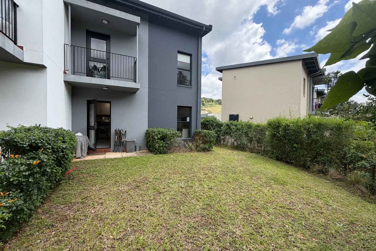 Rear view of a modern townhouse featuring a private garden, paved patio area, and an upper-level balcony with metal railings.