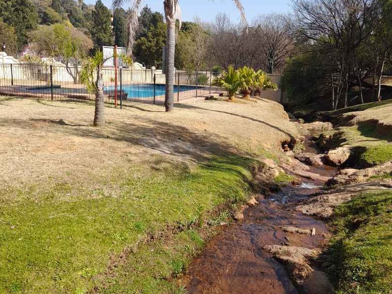A view of the communal swimming pool area featuring secure fencing and landscaped grounds with a natural stream running alongside the property.