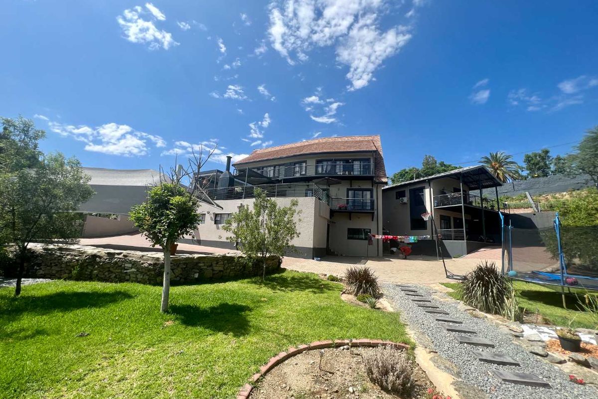 Rear view of a multi-story house featuring a landscaped garden, stone retaining wall, paved driveway, and multiple balconies with metal railings.