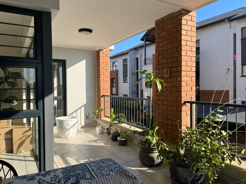 A covered balcony featuring tiled flooring, brick support pillars, and a metal balustrade, offering an outdoor space with views of the apartment complex.