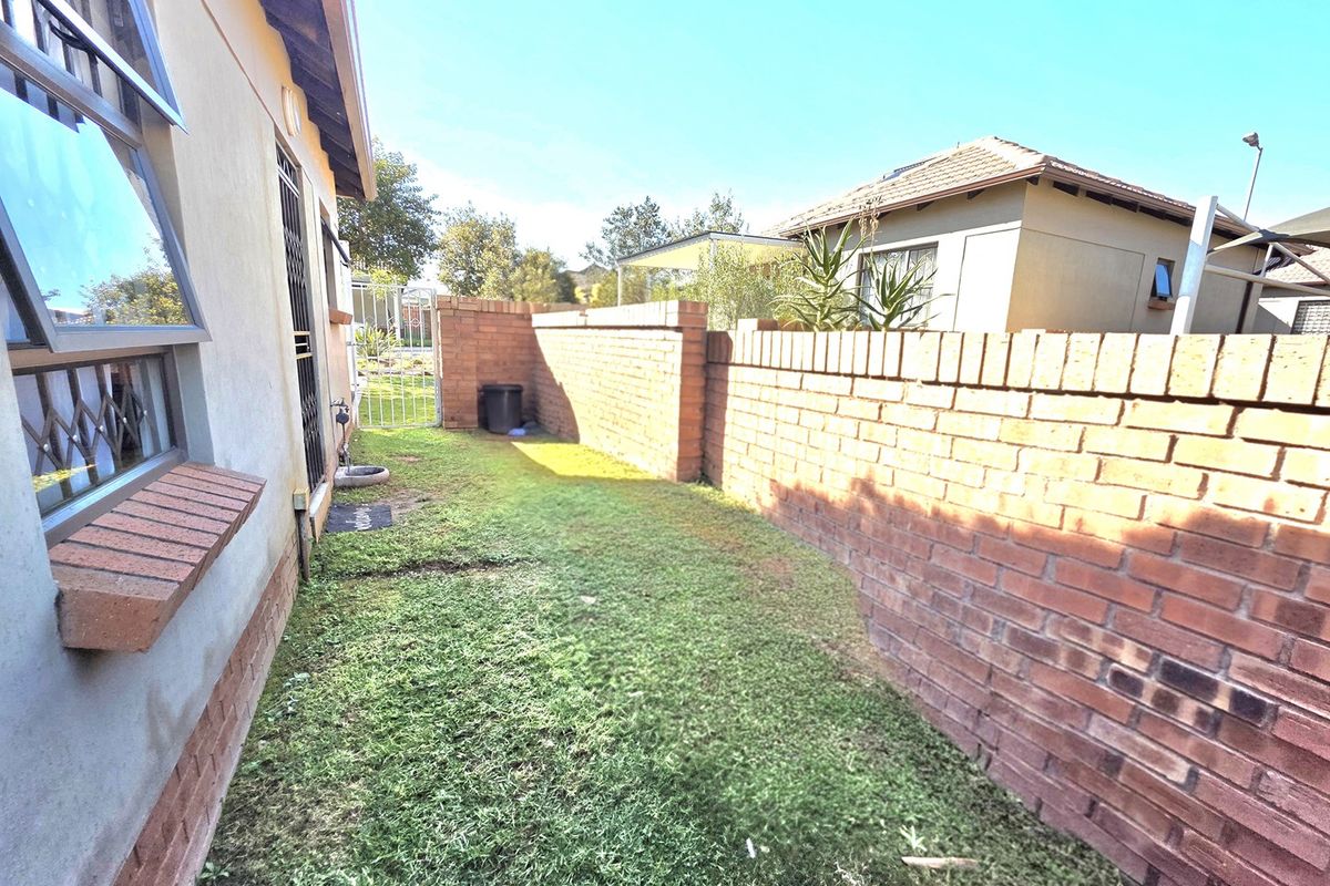 A side garden area of a townhouse featuring a brick boundary wall, a metal security gate, and access to the main building structure.