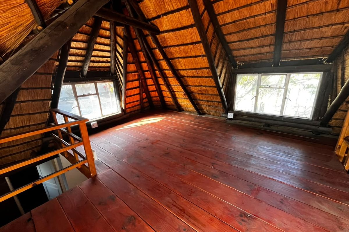 An empty loft-style bedroom featuring exposed timber rafters, a traditional thatched roof, and polished timber flooring with natural light from casement windows.