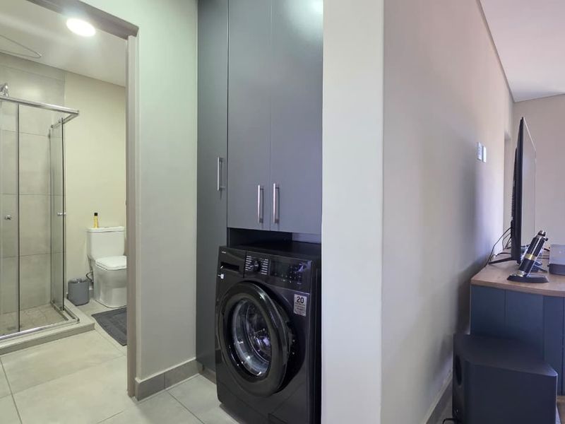 A functional laundry area featuring built-in cabinetry and tiled flooring, conveniently located adjacent to a modern bathroom with a glass shower enclosure.