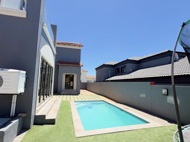 A well-maintained rear courtyard featuring a swimming pool, paved patio area, artificial lawn, and modern grey exterior finishes.