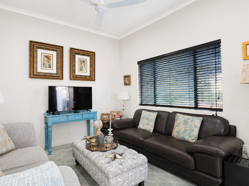A well-maintained living room featuring a ceiling fan, neutral wall finishes, and large windows fitted with venetian blinds.