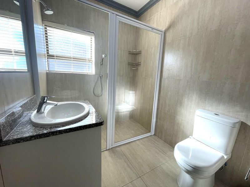 A clean, modern bathroom featuring a granite-topped vanity, a glass-enclosed shower, and a white toilet with neutral floor-to-ceiling wall tiles.