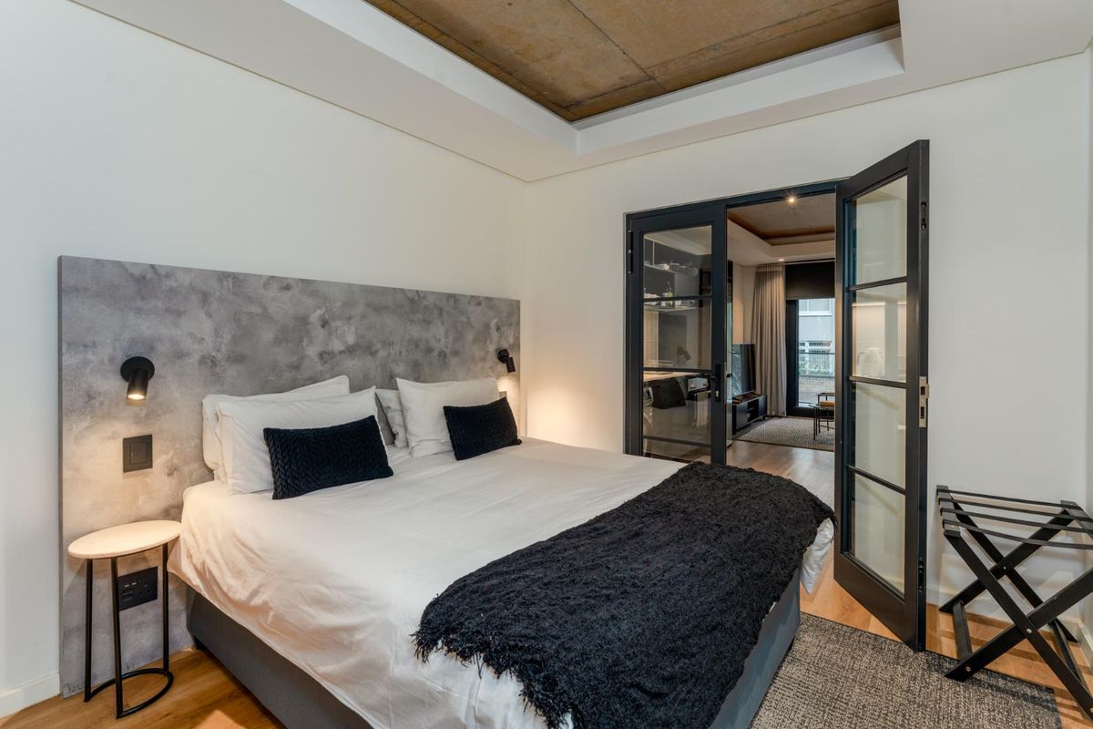 Modern bedroom featuring a unique concrete-finish recessed ceiling, integrated headboard with wall-mounted lighting, and elegant glass-paneled double doors leading to an adjacent living space.