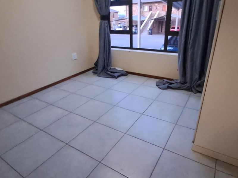 A bright, empty bedroom featuring neutral tiled flooring, a built-in cupboard, and a large window providing natural light and views of the complex.