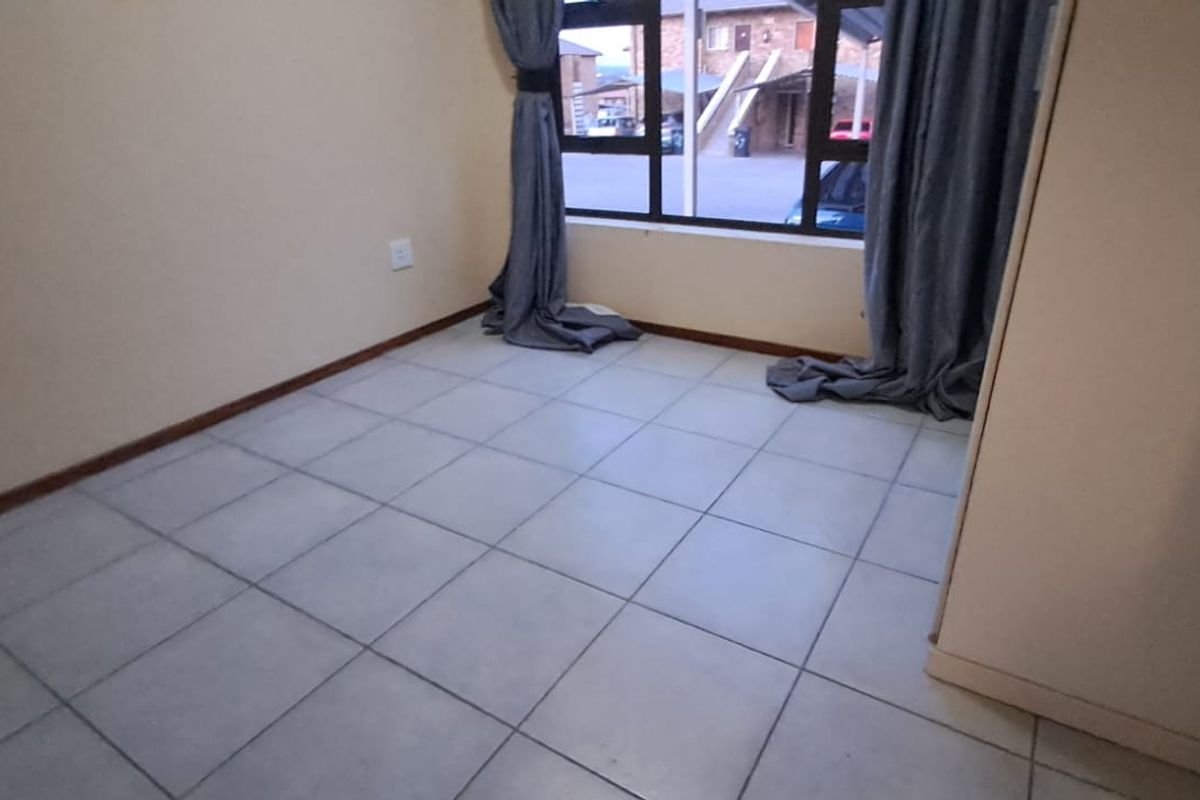 A bright, empty bedroom featuring neutral tiled flooring, a built-in cupboard, and a large window providing natural light and views of the complex.