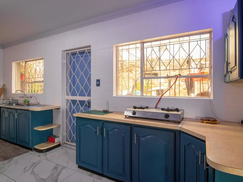 A functional kitchen featuring blue cabinetry, laminate countertops, a gas hob, and tiled finishes, with direct access to the exterior via a security door.