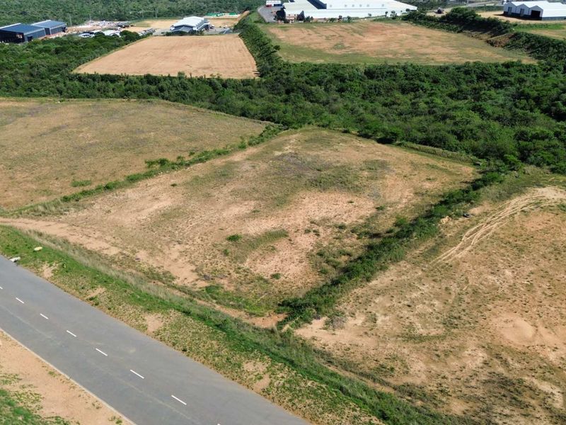 Aerial view of an undeveloped industrial land plot adjacent to a paved road, featuring natural vegetation and surrounding industrial infrastructure.