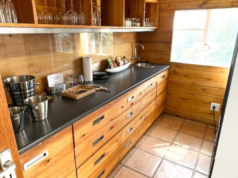 A functional scullery area featuring timber wall cladding, extensive wooden cabinetry, a dark laminate countertop, and a stainless steel sink with natural light.