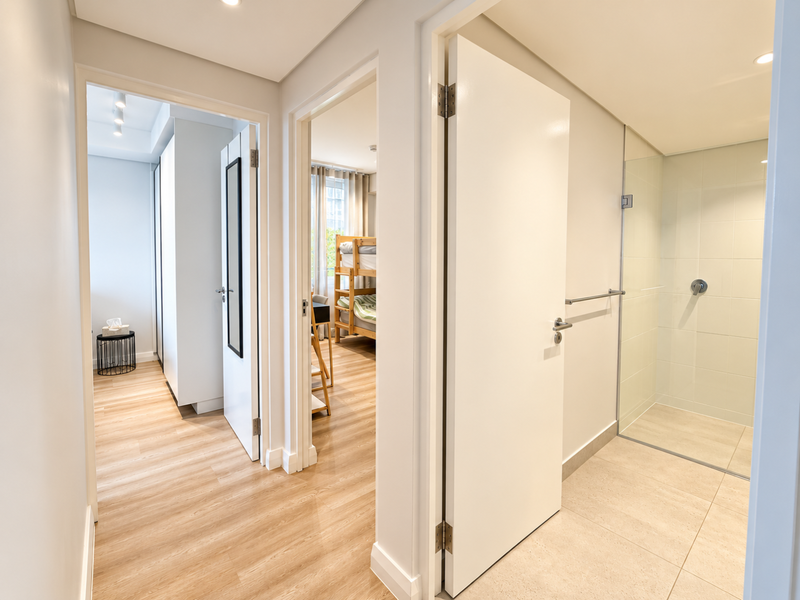A bright, modern hallway featuring laminate flooring, recessed lighting, and access to a bedroom and a bathroom with a glass-enclosed shower.