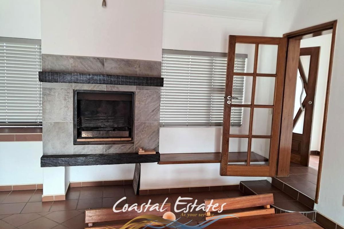 A bright living room featuring a prominent built-in fireplace with stone cladding and a timber mantel, complemented by tiled flooring and timber-framed glass doors.