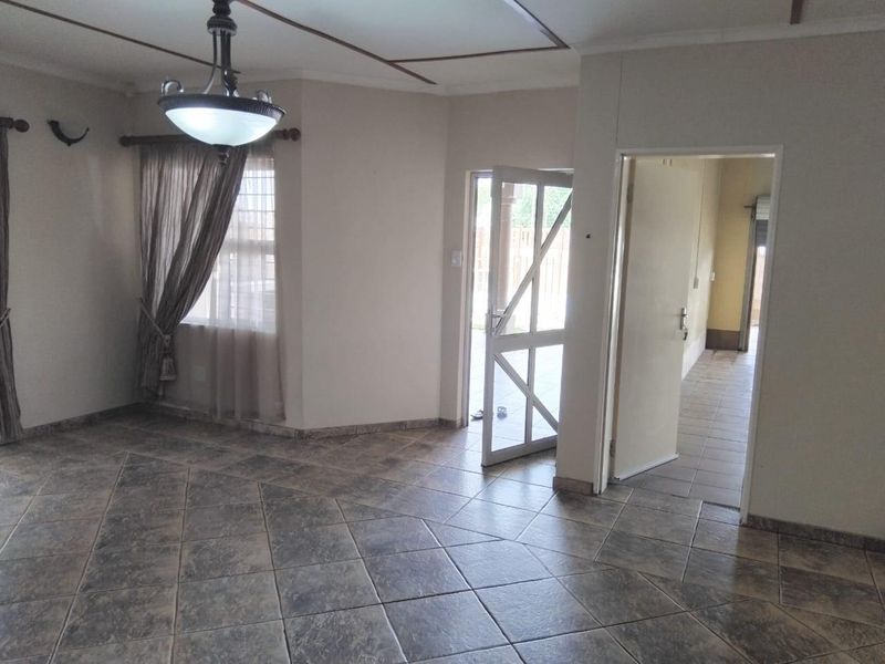 An empty living room featuring tiled flooring, a central pendant light fixture, and access to an adjacent hallway and exterior area via a security gate.