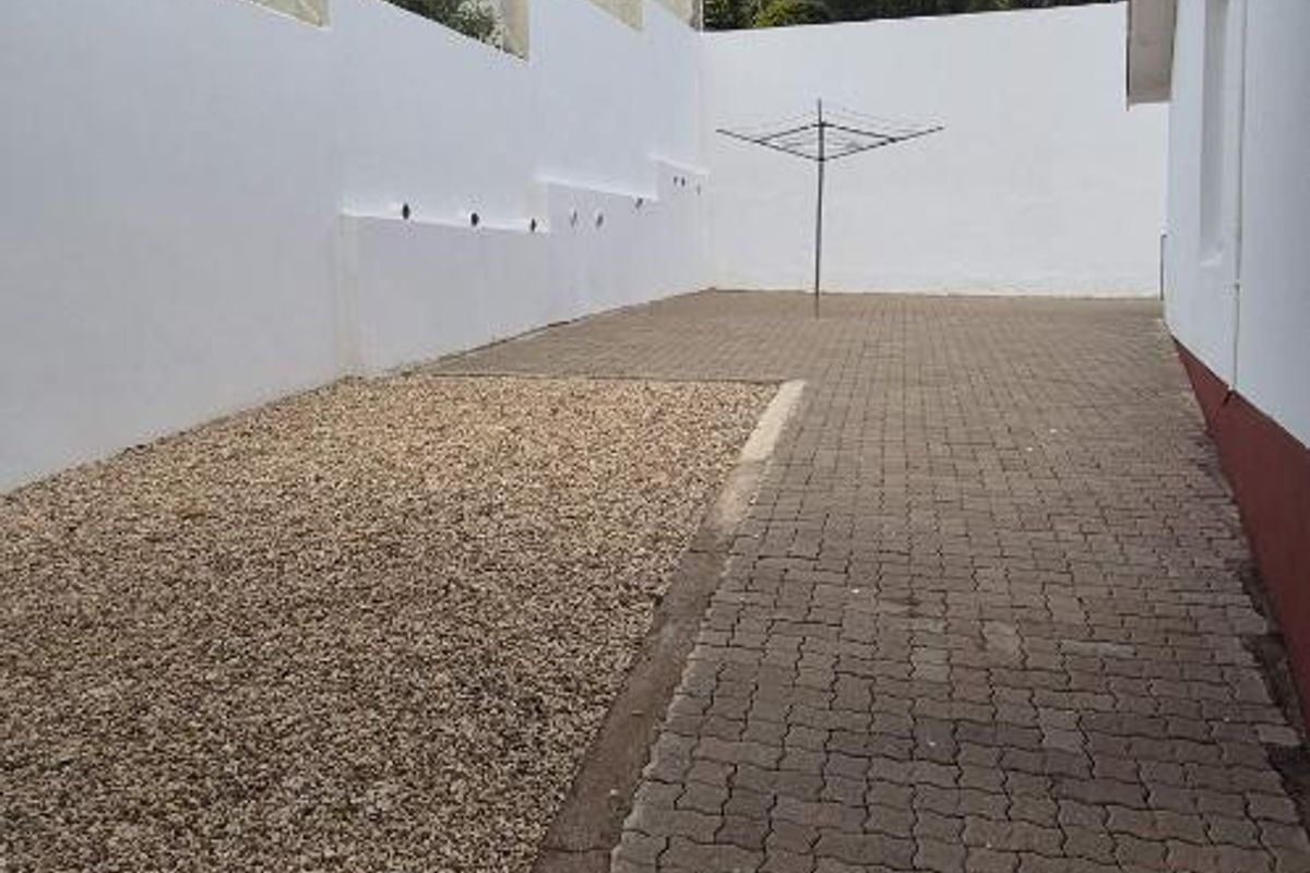 A paved side yard area featuring a gravel section, high perimeter boundary walls, and an external wall-mounted air conditioning unit.