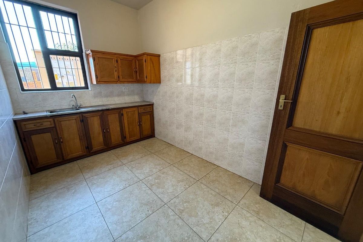 A functional kitchen area featuring wooden cabinetry, tiled splashbacks, a stainless steel sink, and tiled flooring with a security-barred window.