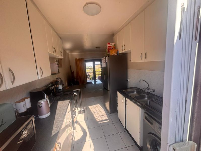 A functional galley-style kitchen featuring white cabinetry, tiled flooring, double basin sink, and built-in cooking appliances, leading towards an open living area.