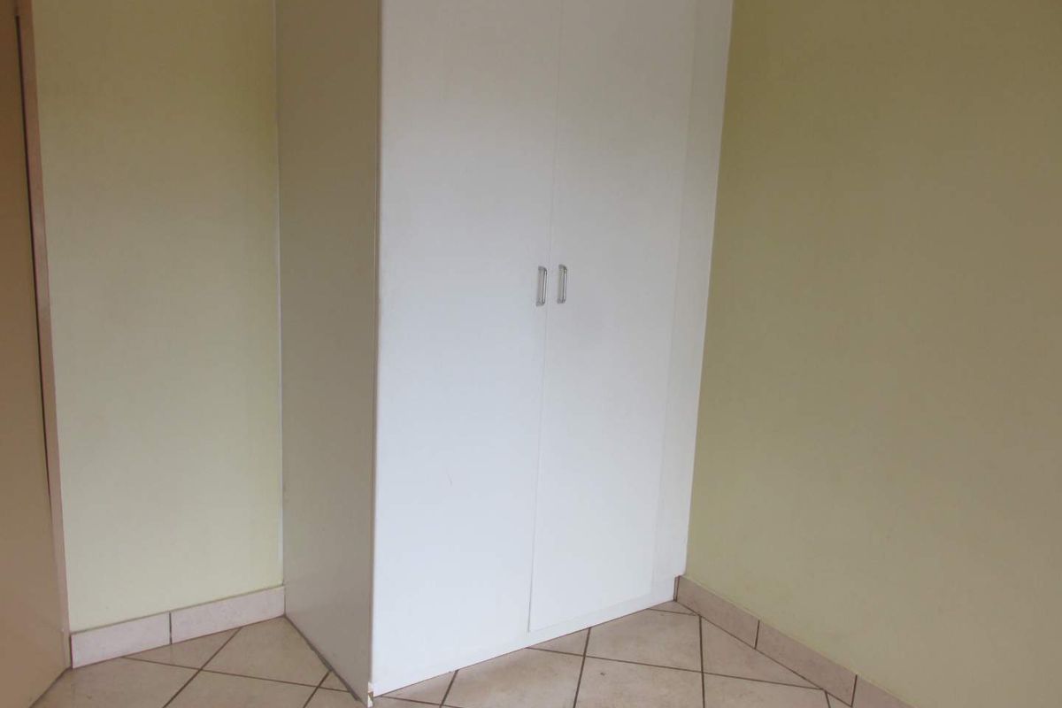A simple bedroom corner featuring a white built-in cupboard and neutral tiled flooring with matching tiled skirting.