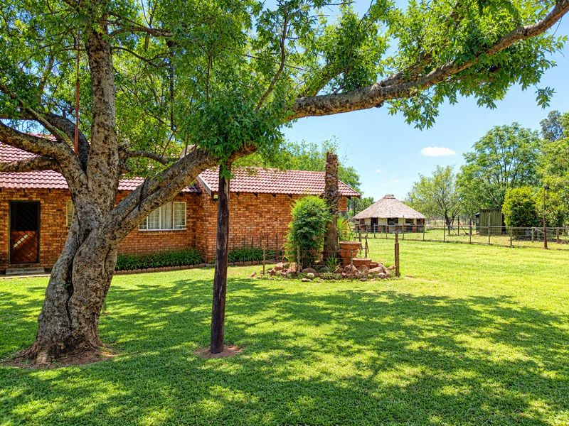 A spacious rear garden featuring a face brick dwelling with a tiled roof, mature trees, and a separate thatched lapa structure on the property.