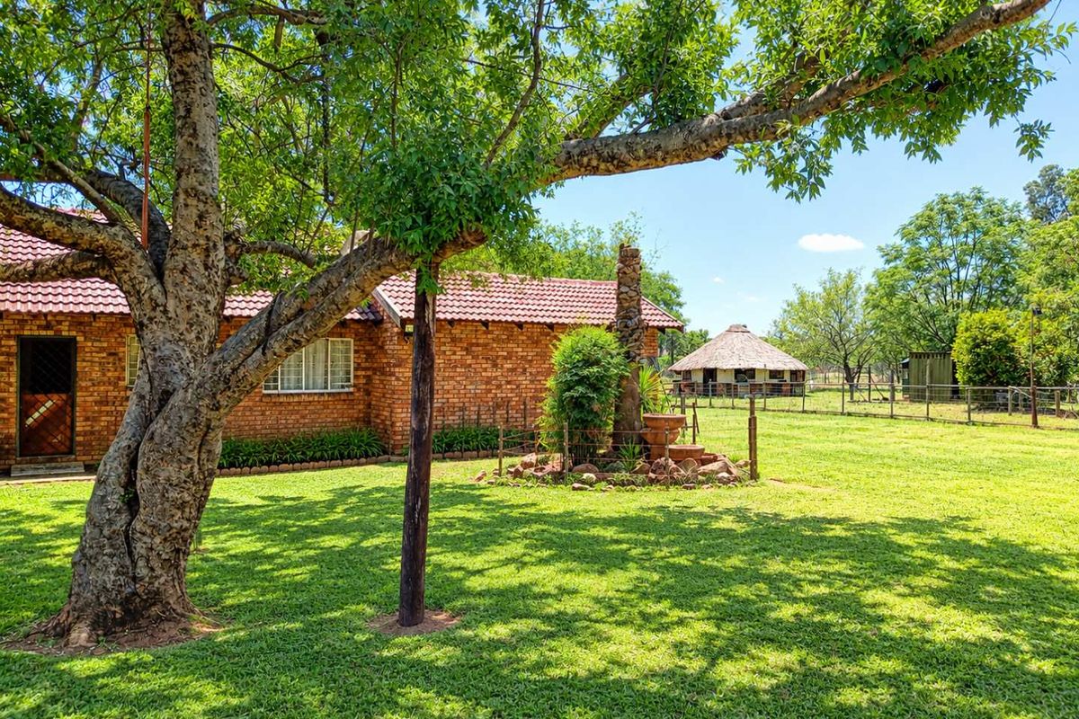 A spacious rear garden featuring a face brick dwelling with a tiled roof, mature trees, and a separate thatched lapa structure on the property.