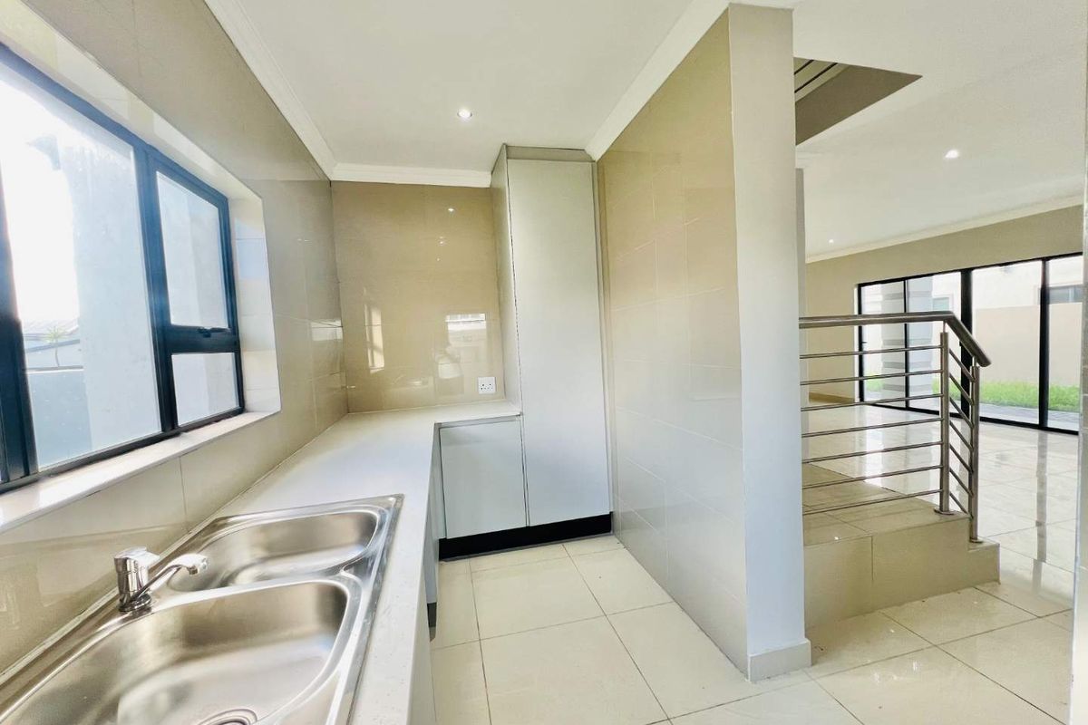 Modern kitchen area featuring sleek cabinetry, stone countertops, a double stainless steel sink, and tiled flooring, adjacent to a staircase with stainless steel railings.
