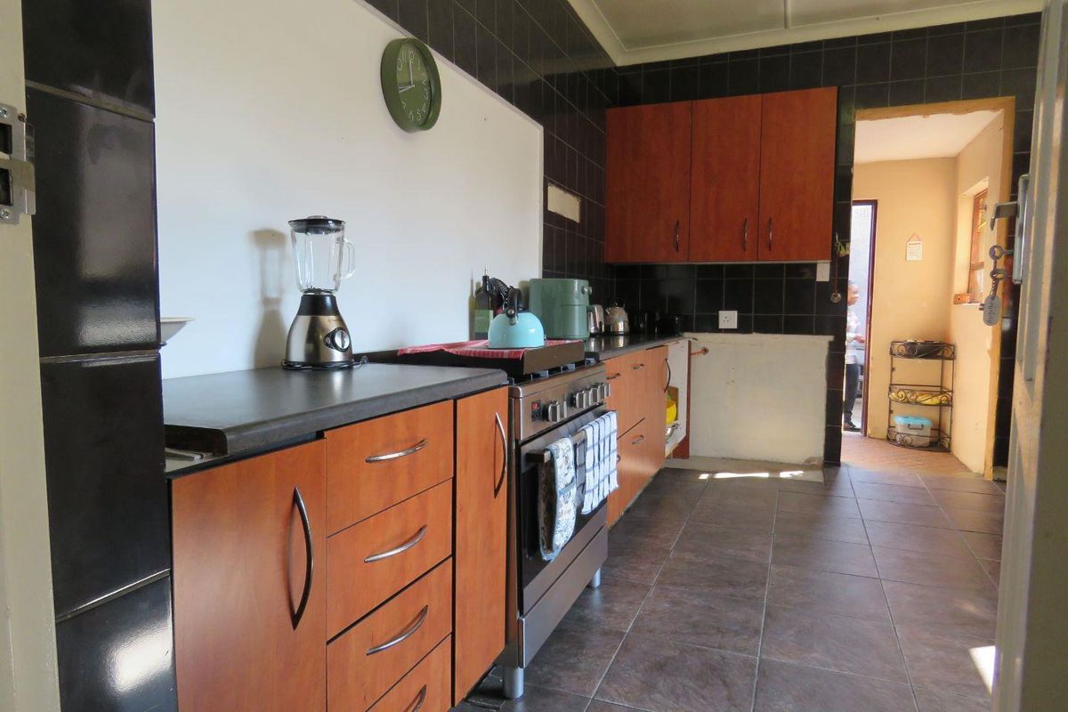 A functional kitchen featuring wood-finish cabinetry, dark tiled splashbacks, tiled flooring, and a freestanding stove, with an adjacent doorway leading to a utility area.
