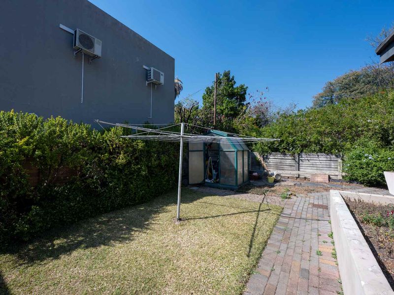 A rear garden area featuring a lawn, paved walkway, rotary clothesline, and a small greenhouse, bordered by established hedges and a timber fence.