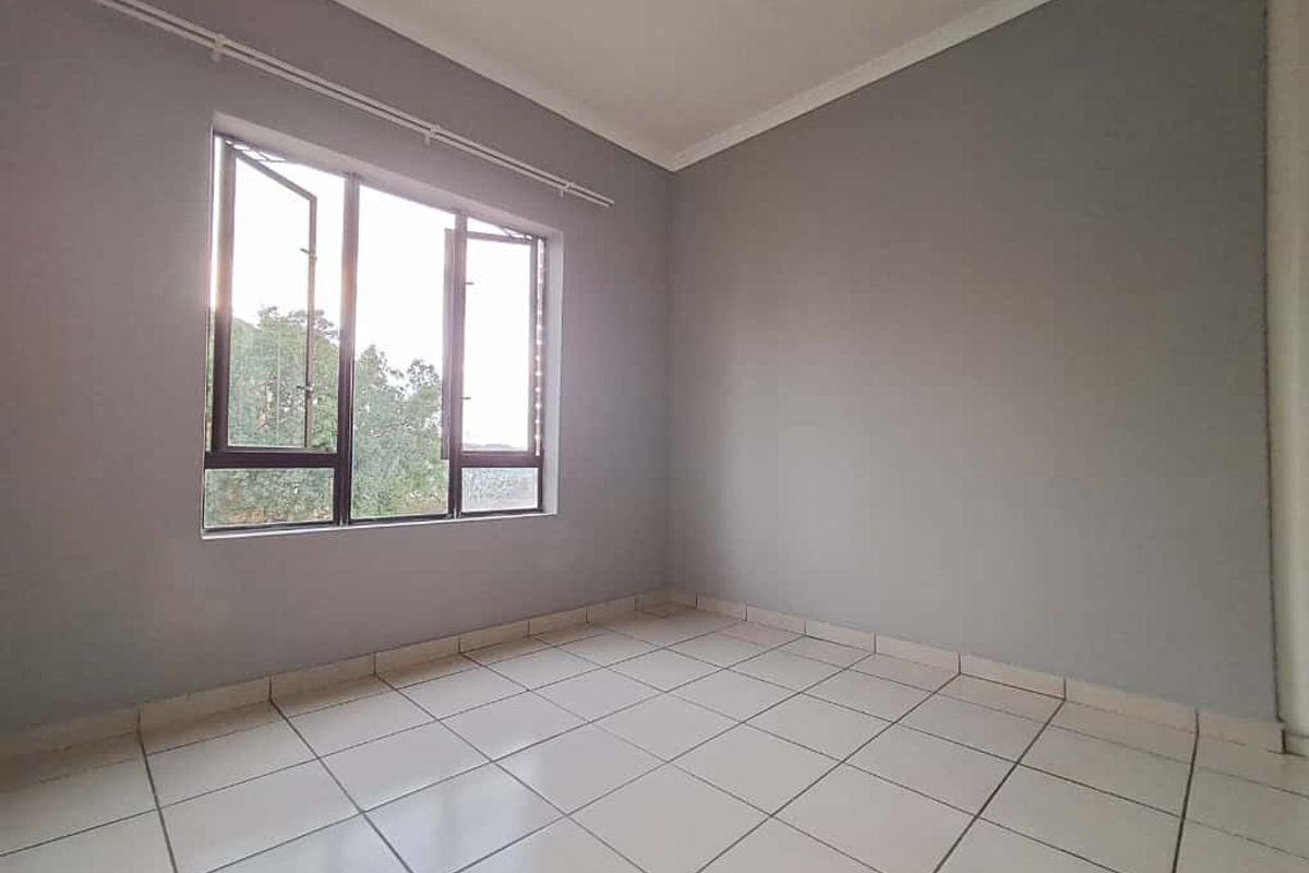 A bright, empty bedroom featuring neutral grey walls, tiled flooring, and a large window providing natural light.