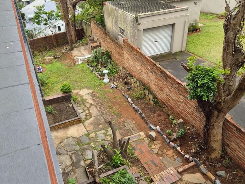 Elevated view of a rear garden featuring a brick boundary wall, paved pathways, stone patio, built-in braai, and a detached garage.