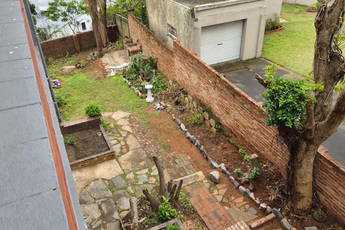 Elevated view of a rear garden featuring a brick boundary wall, paved pathways, stone patio, built-in braai, and a detached garage.