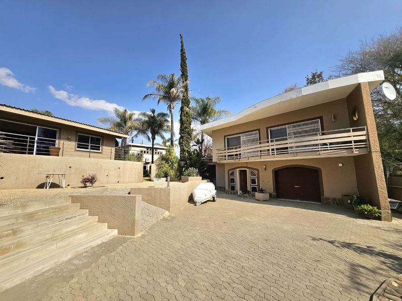 Rear exterior view of the property featuring a paved driveway, multi-level terraced garden with retaining walls, and a balcony overlooking the yard.