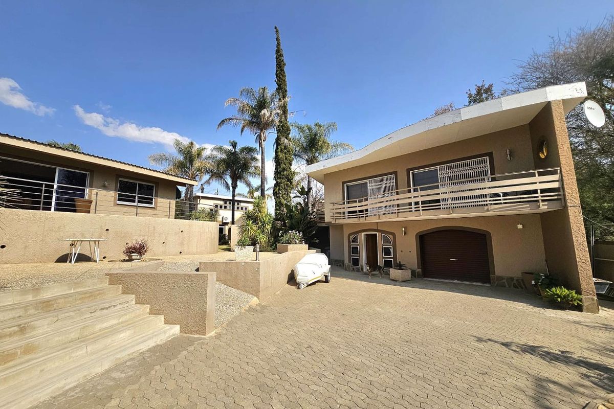 Rear exterior view of the property featuring a paved driveway, multi-level terraced garden with retaining walls, and a balcony overlooking the yard.