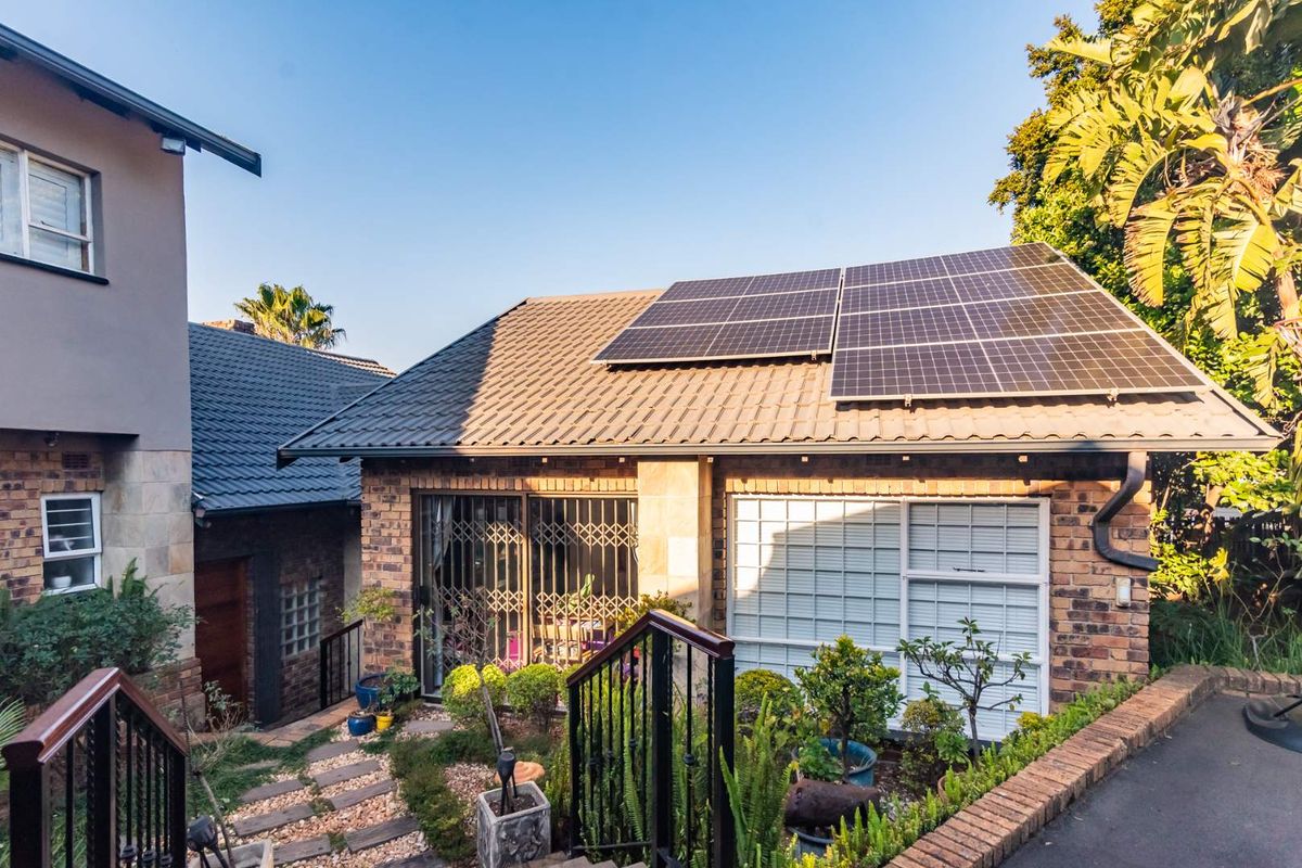 Rear view of the property featuring a brick facade, tiled roof with installed solar panels, and a landscaped garden area with paved walkway.