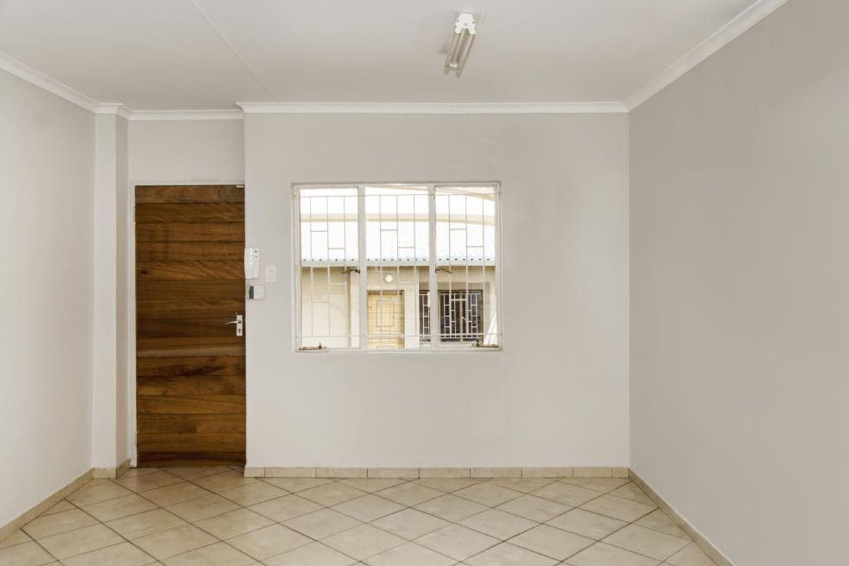 A bright, empty living area featuring neutral wall finishes, tiled flooring, a wooden entrance door, and a window with security bars.