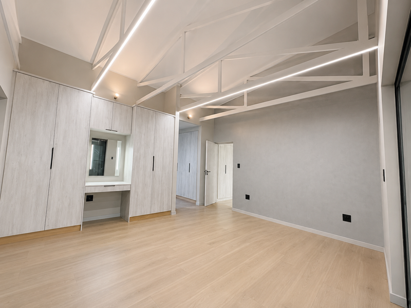 Spacious bedroom featuring light wood-look laminate flooring, extensive built-in cabinetry with a vanity, and modern exposed white roof trusses with integrated linear lighting.
