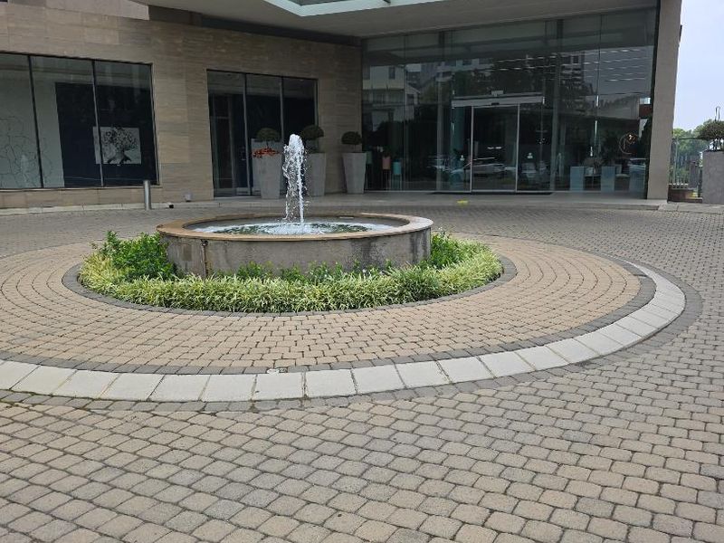 The front entrance of the apartment building features a paved circular driveway, a central water fountain, and a modern glass facade with a covered canopy.