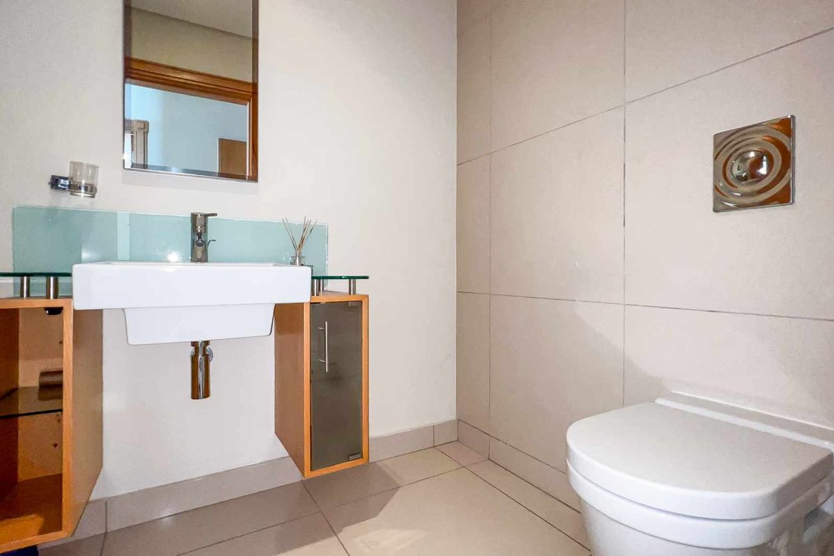 A modern bathroom featuring a wall-mounted vanity with a glass countertop, a wall-hung toilet with a chrome flush plate, and neutral floor-to-ceiling tiling.