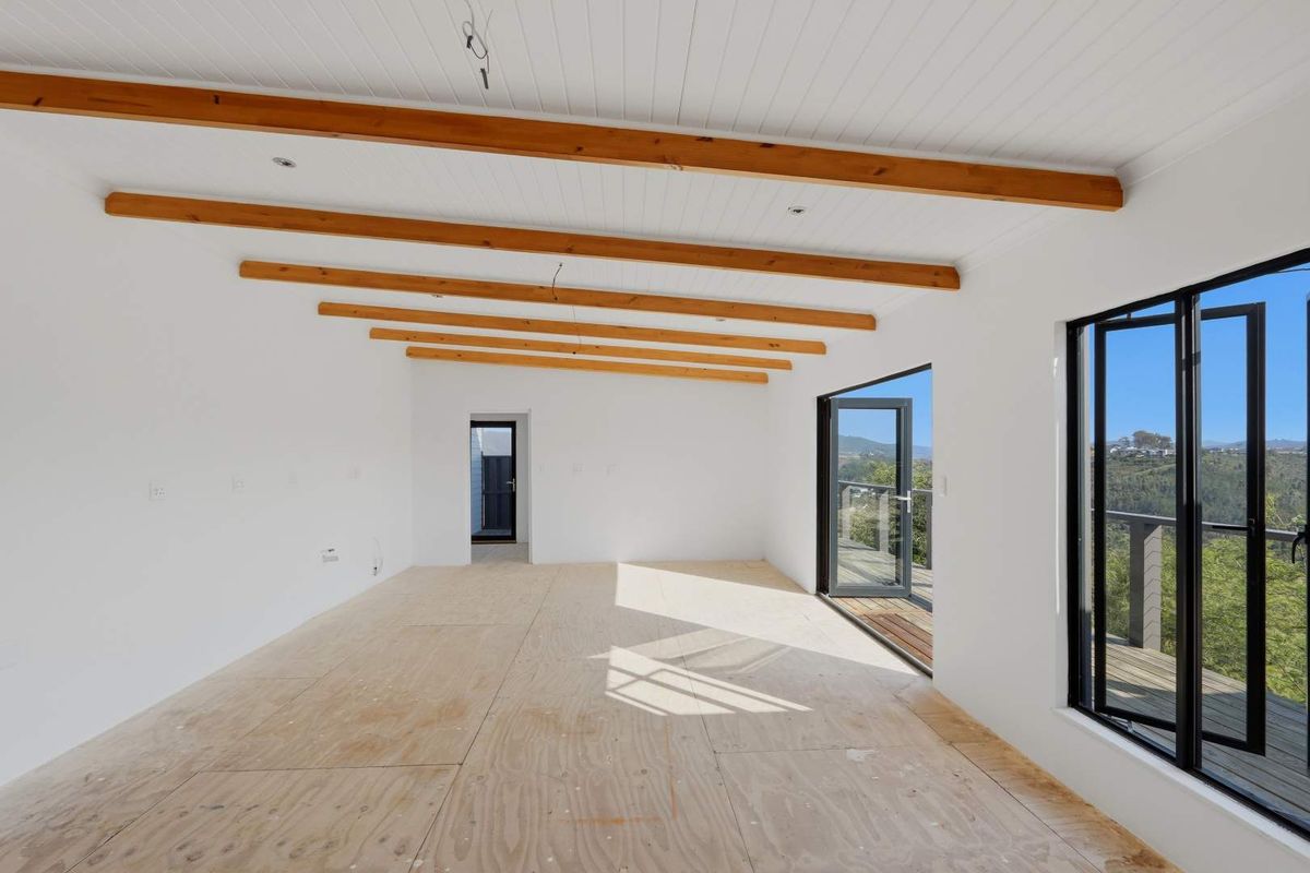 An empty, unfinished living space featuring exposed timber ceiling beams, large windows, and direct access to an outdoor deck with scenic views.