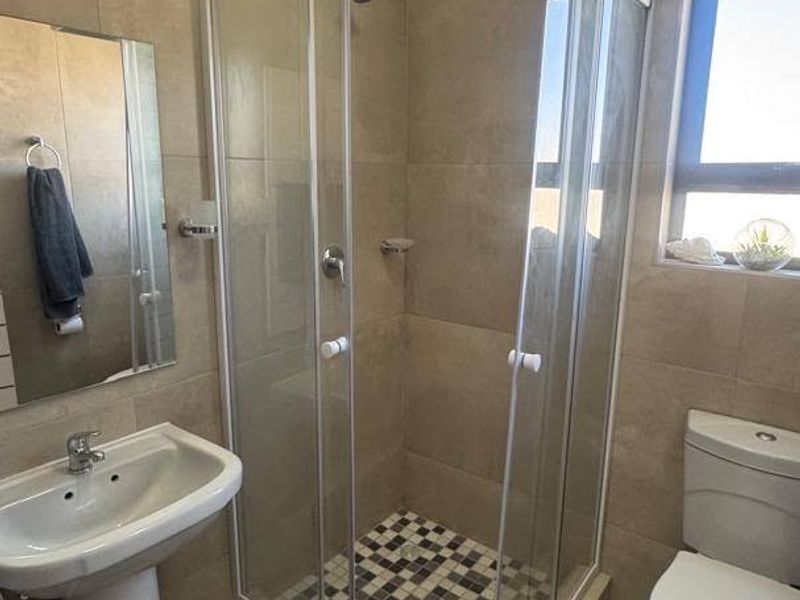 A functional bathroom featuring a pedestal basin, a glass-enclosed shower with mosaic floor tiling, and a standard toilet with neutral wall tiles.