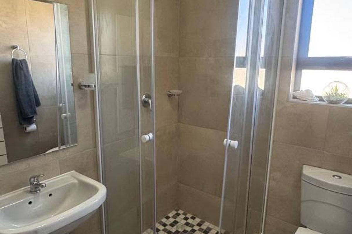 A functional bathroom featuring a pedestal basin, a glass-enclosed shower with mosaic floor tiling, and a standard toilet with neutral wall tiles.