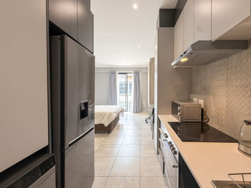 Modern open-plan apartment featuring a sleek kitchen with built-in appliances and tiled flooring, seamlessly flowing into the bedroom area with balcony access.