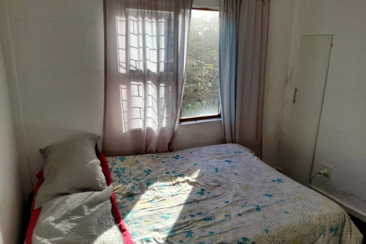 A compact bedroom featuring a window with natural light, built-in cupboard, and basic wall finishes.