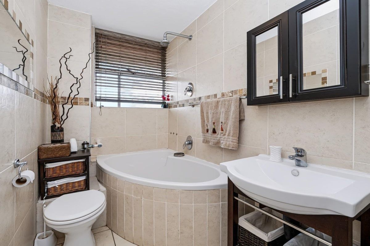 A functional bathroom featuring a corner bathtub, wall-mounted shower, vanity basin, and neutral ceramic tiling throughout.
