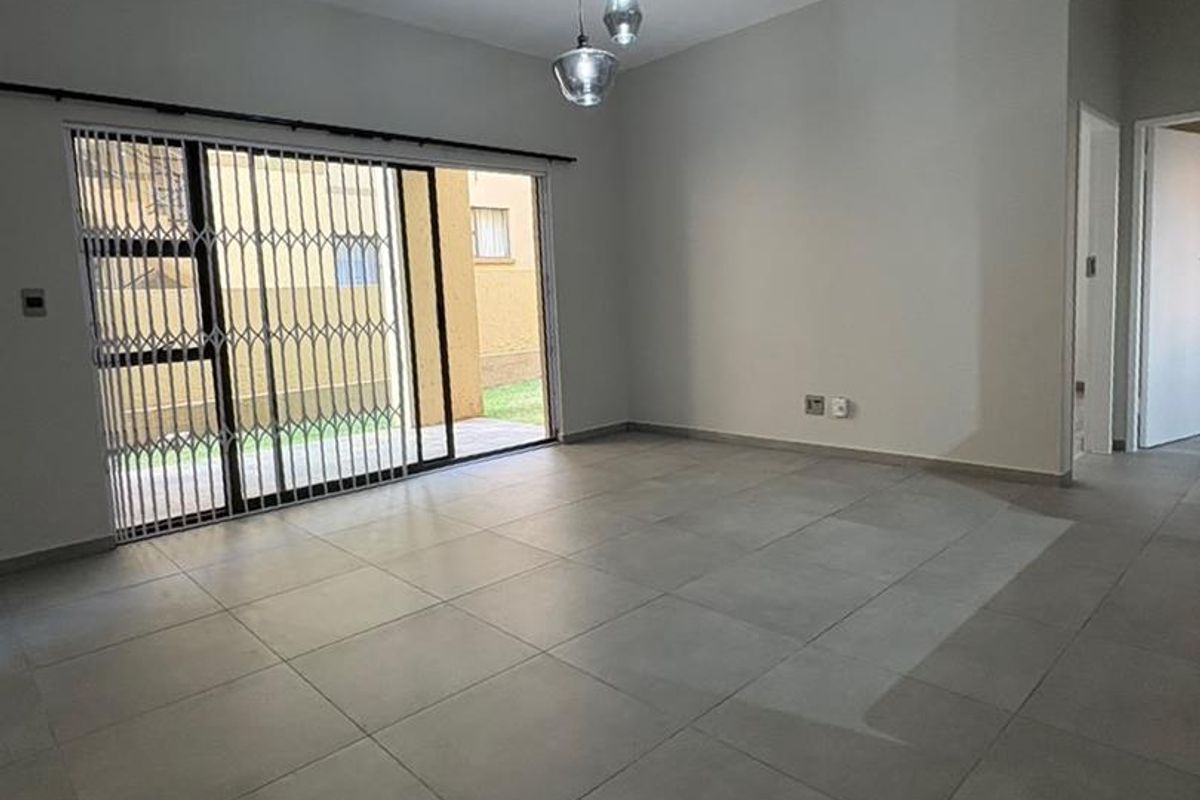 A bright, empty living room featuring large format floor tiles, a modern pendant light, and a sliding glass door with a security gate leading outside.